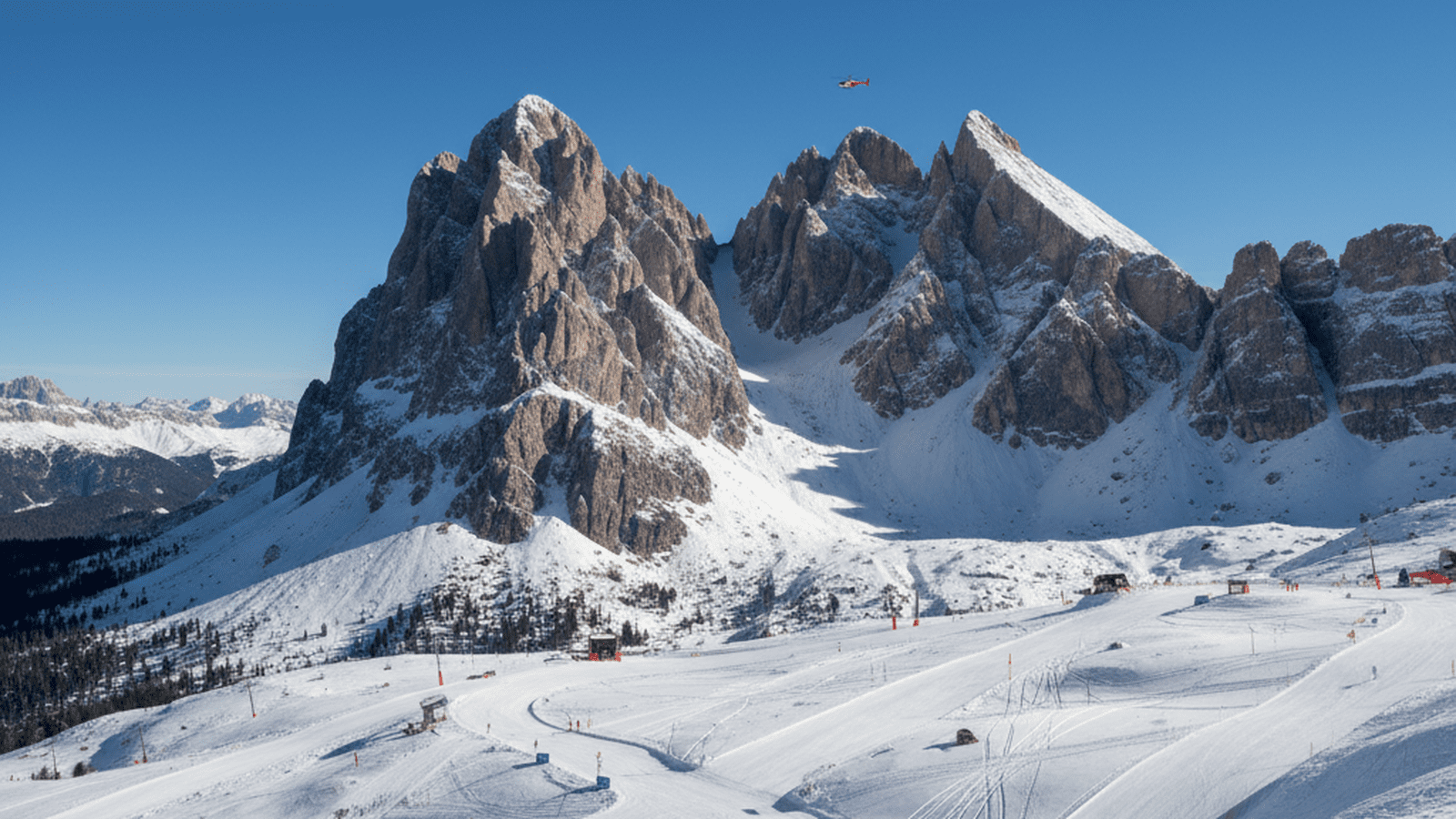 A high-altitude view of a steep Olympic ski course in the Italian Dolomites with a rescue helicopter in the distance.