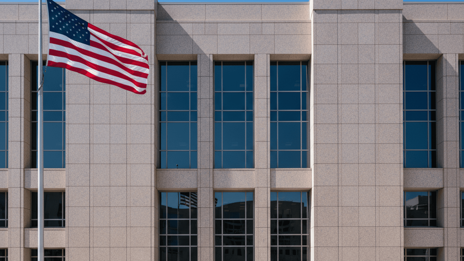 A large American flag flies in front of a modern, imposing federal government building under a clear blue sky.
