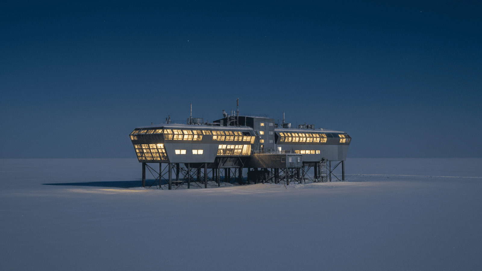 A high-tech research station stands on stilts amidst a vast, frozen Antarctic landscape under a dark blue sky.