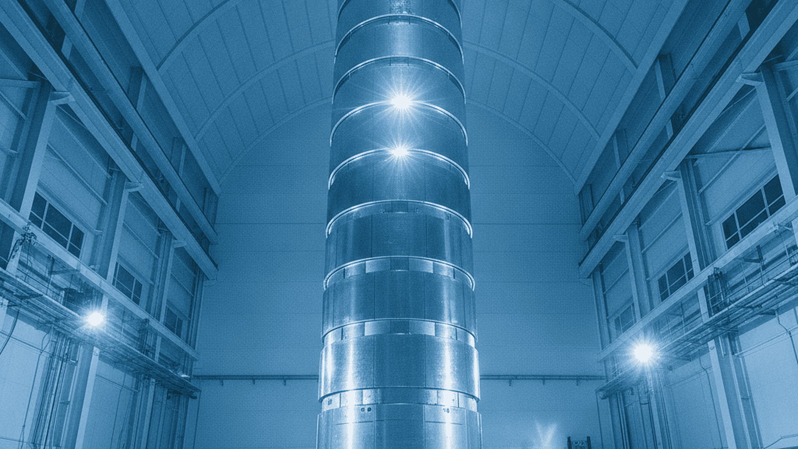 A massive silver metallic tower being tested inside a large aerospace assembly facility with engineers at its base.