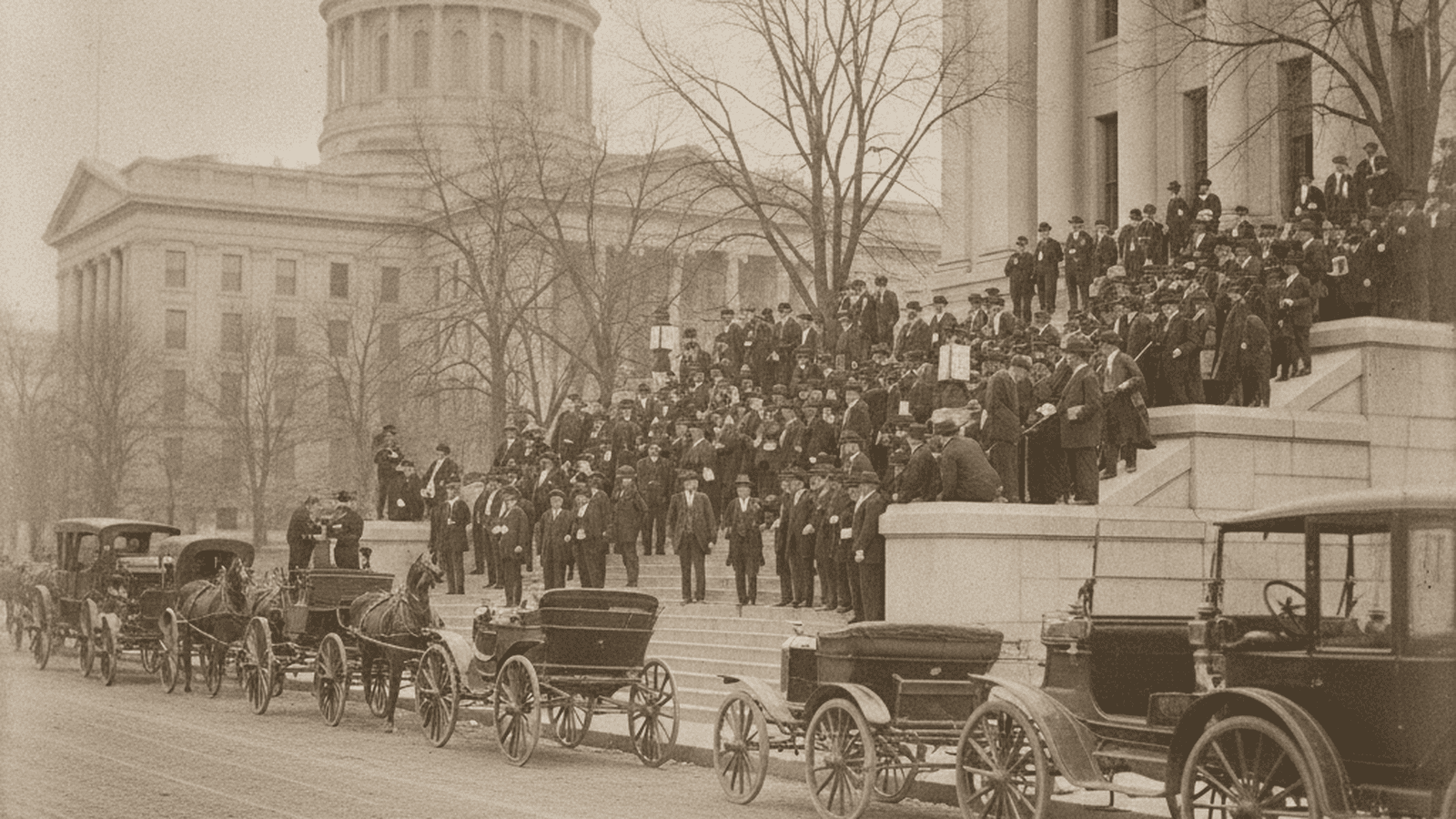 A sepia-toned 1912 photograph of the Kansas State Capitol with men in period-accurate suits and early automobiles.