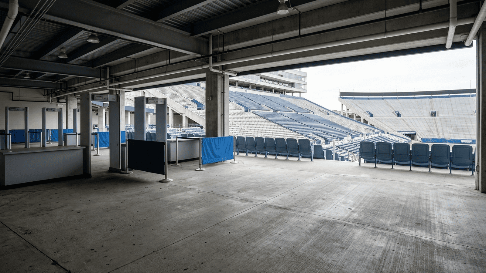 3e8ac91d381e1a43e7960e22540c795e0d20656a627a972135f8a3680a14ed28 An empty football stadium concourse looks toward the seating area beyond a security checkpoint.