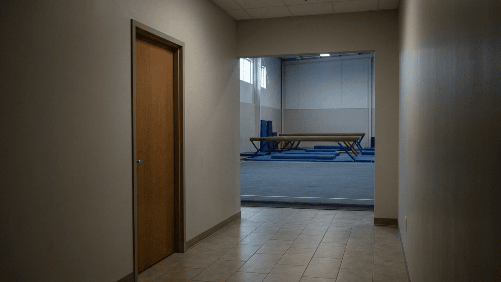 An empty gymnastics facility hallway with a closed bathroom door and training equipment in the distance.