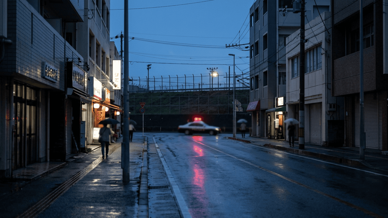 7737cdc75a1732e466bbbaed51e3f86dbbadfcf73aa4ac60fc9ec36f4c2891a9 A quiet Okinawa street at dusk with police lights in the distance and military-base fencing nearby.