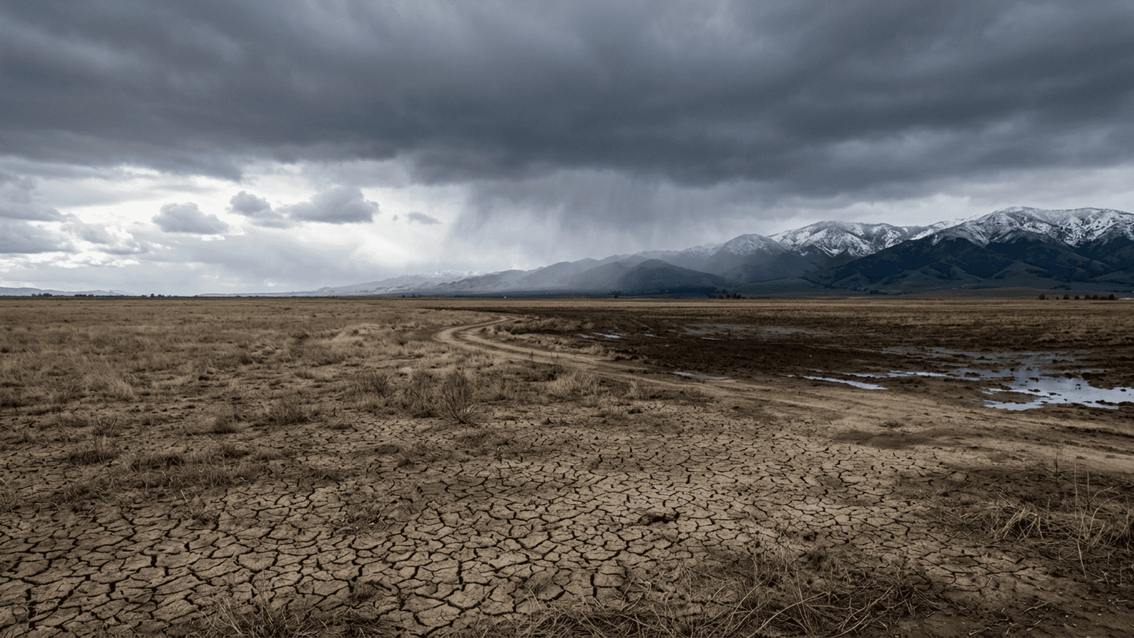 A winter storm approaches a landscape where dry cracked ground meets rain-darkened soil.