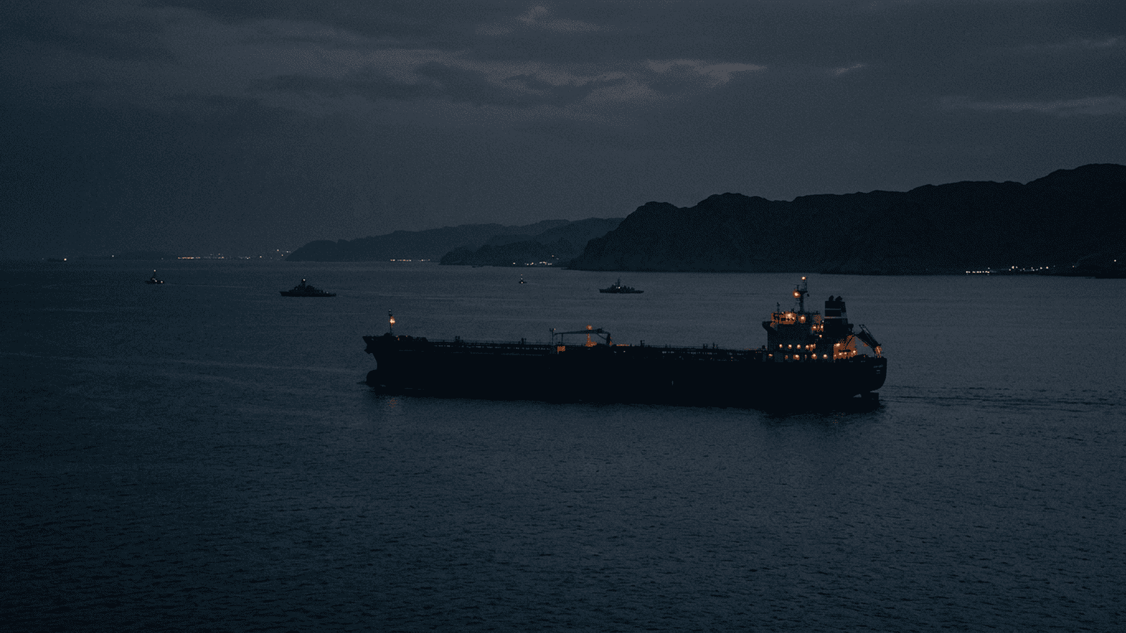 An oil tanker travels through the Strait of Hormuz at night with naval vessels nearby.
