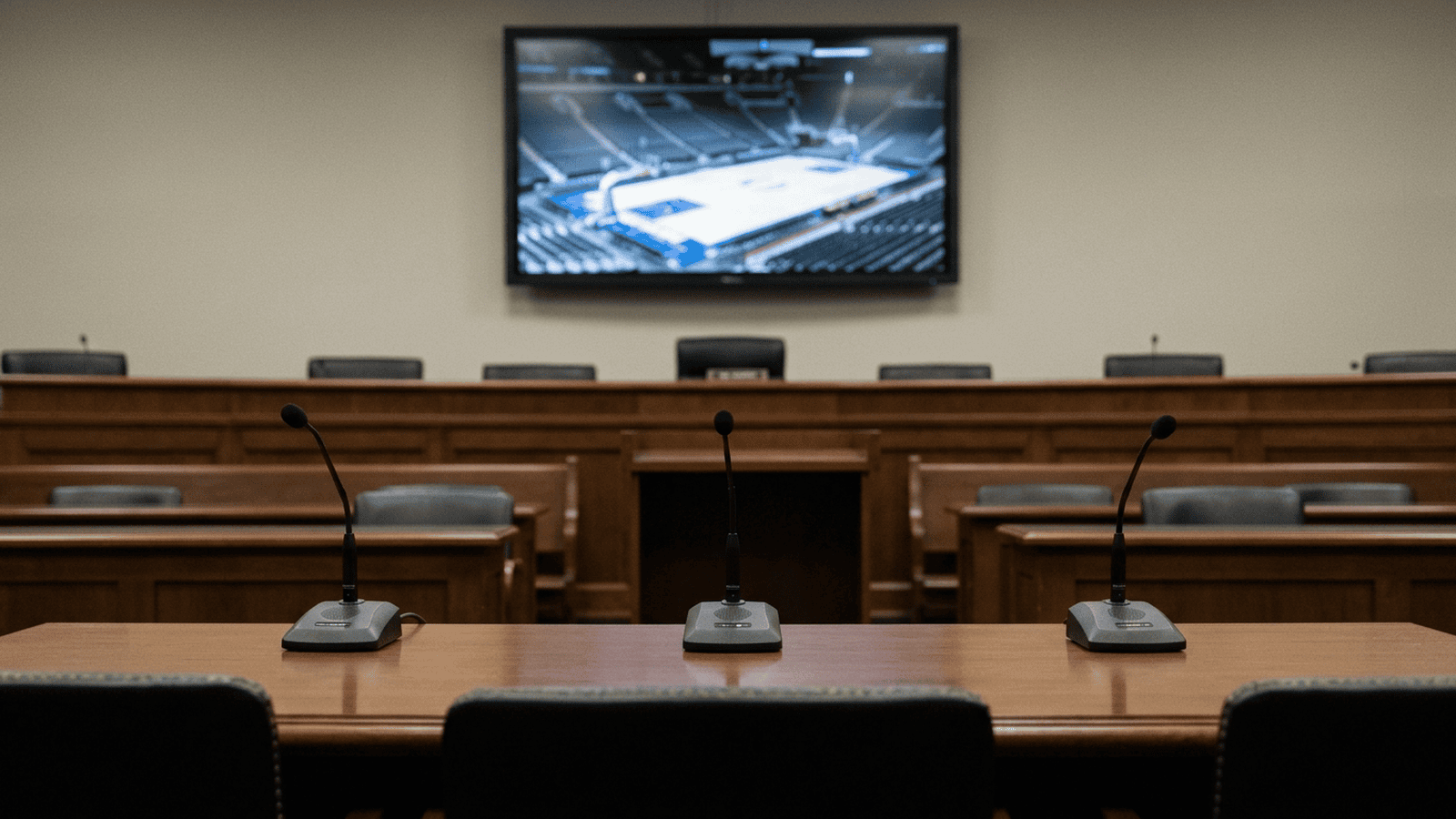 An empty Senate hearing room with microphones and a blurred basketball arena displayed on a screen.