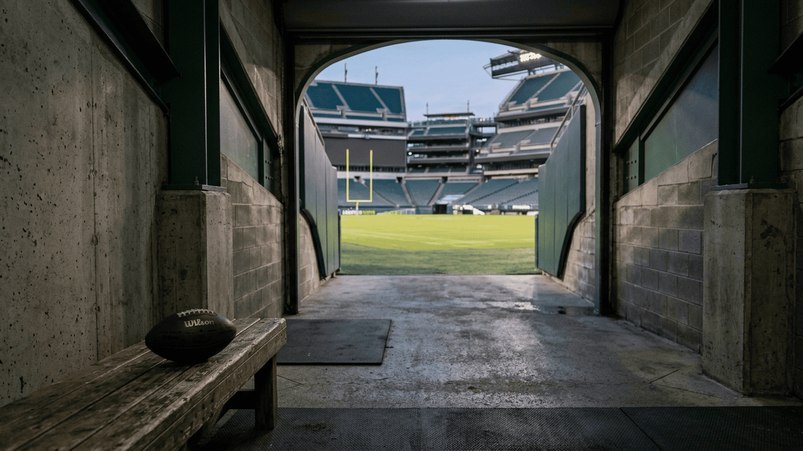 d1ba2127828f525ce7e2705bc246e81a47fb4afc8ab3e930d1dc822ba3d8512b An empty football stadium tunnel opens toward a lit field with a football resting on a bench.