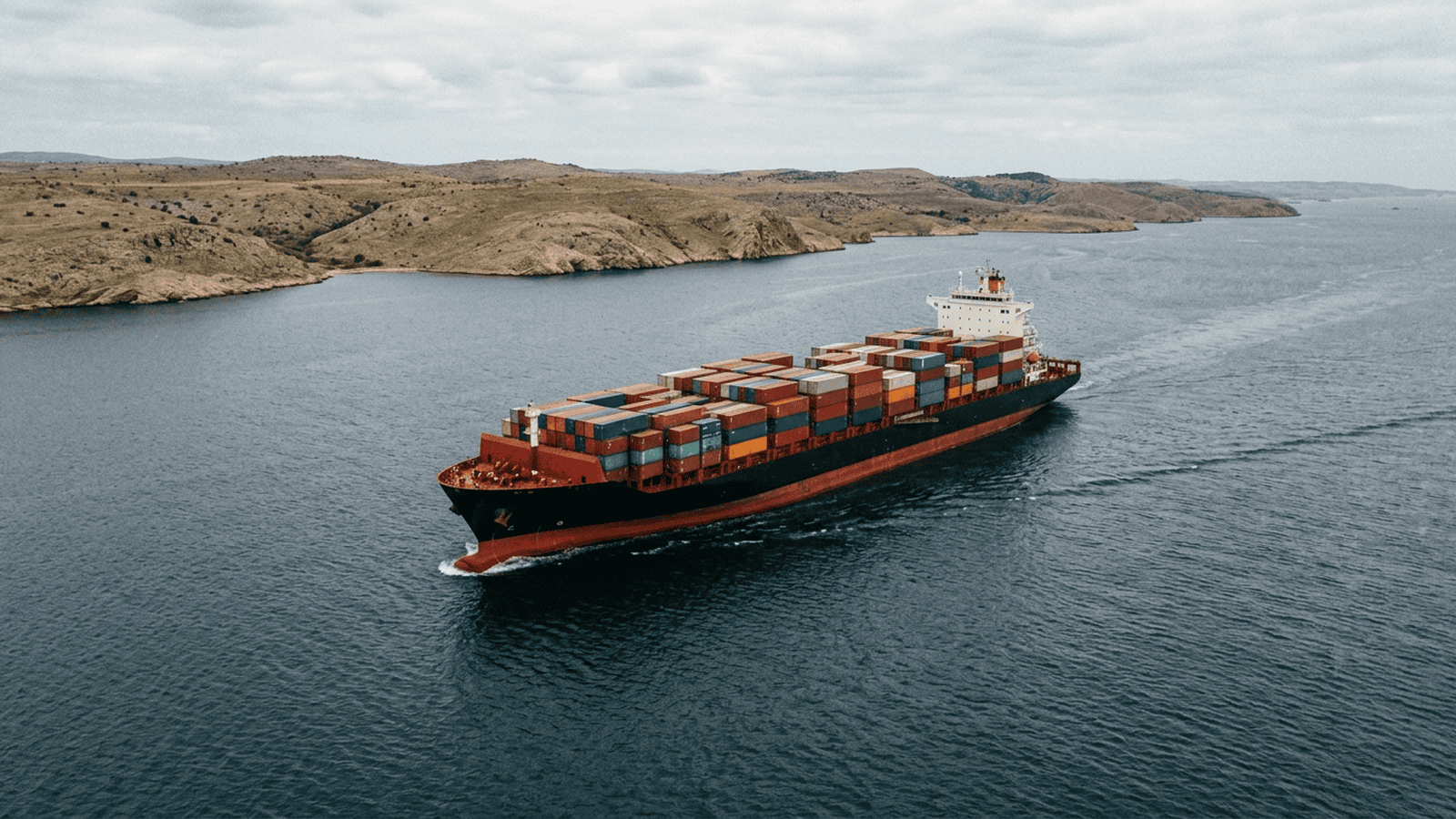 A cargo ship travels through a broad waterway under a cloudy sky.