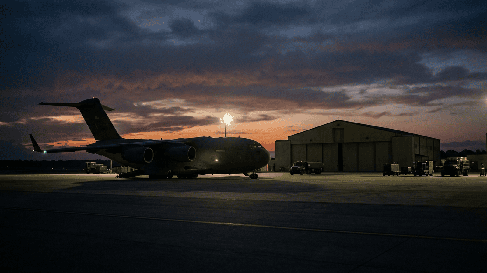 A military aircraft sits on a runway near a hangar at dusk.